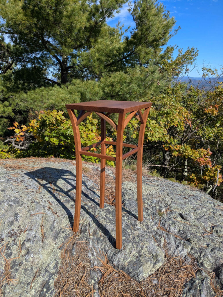 An elegant, small end table with curved legs on a rock at the summit of Jug End, in front of some evergreen and oak trees in the autumn. There is a filtered view through the trees to the mountains in the distance.